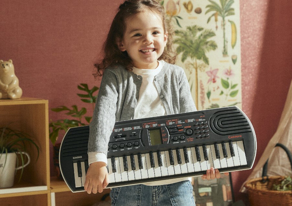 Dziecko trzyma czarny keyboard marki Casio z widocznymi klawiszami i przyciskami. W tle znajduje się roślinny plakat i drewniana półka z dekoracjami.