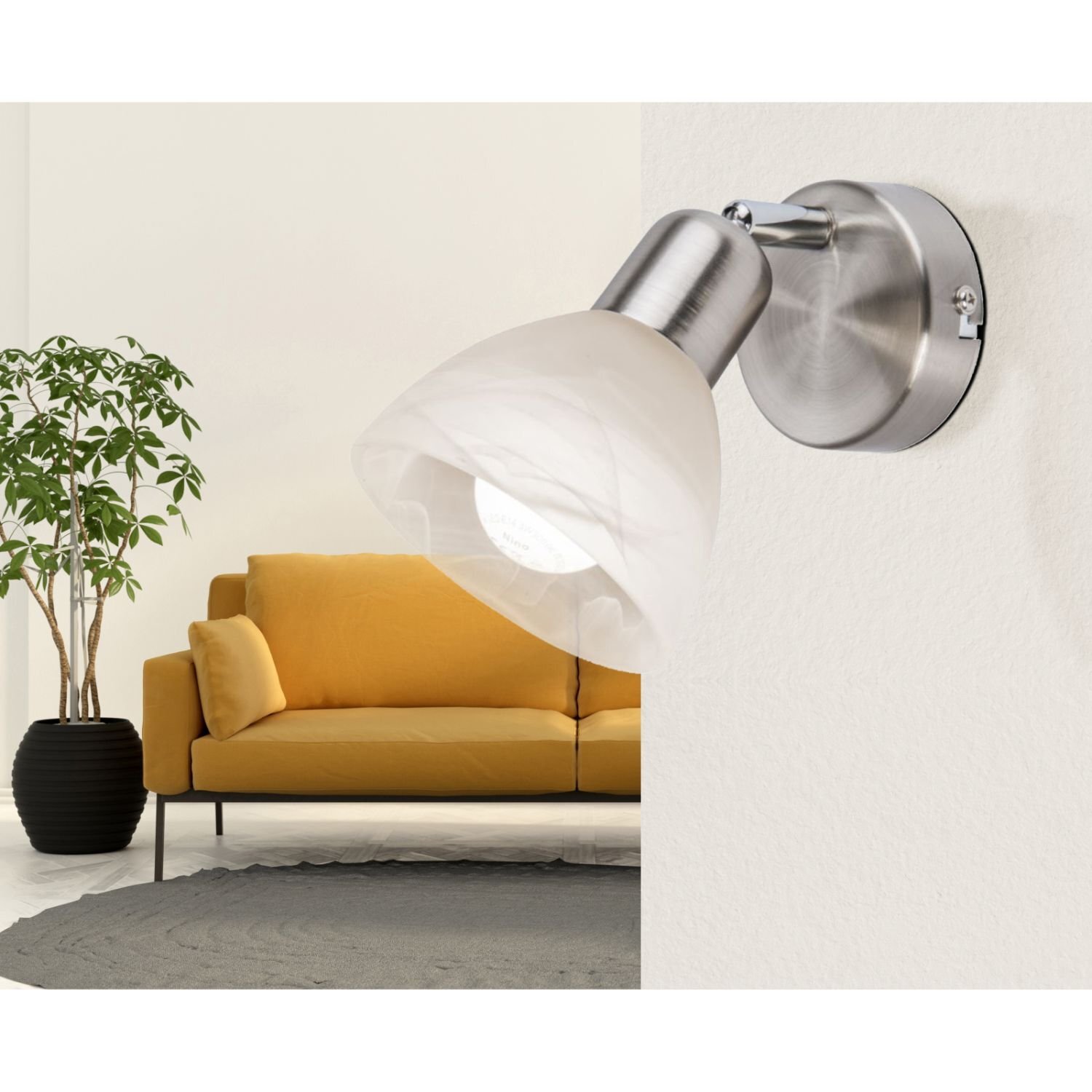 Na obrazie widoczny jest reflektor spot zamontowany na ścianie, wykonany z metalu i szkła. W tle znajduje się żółta sofa oraz roślina w doniczce.