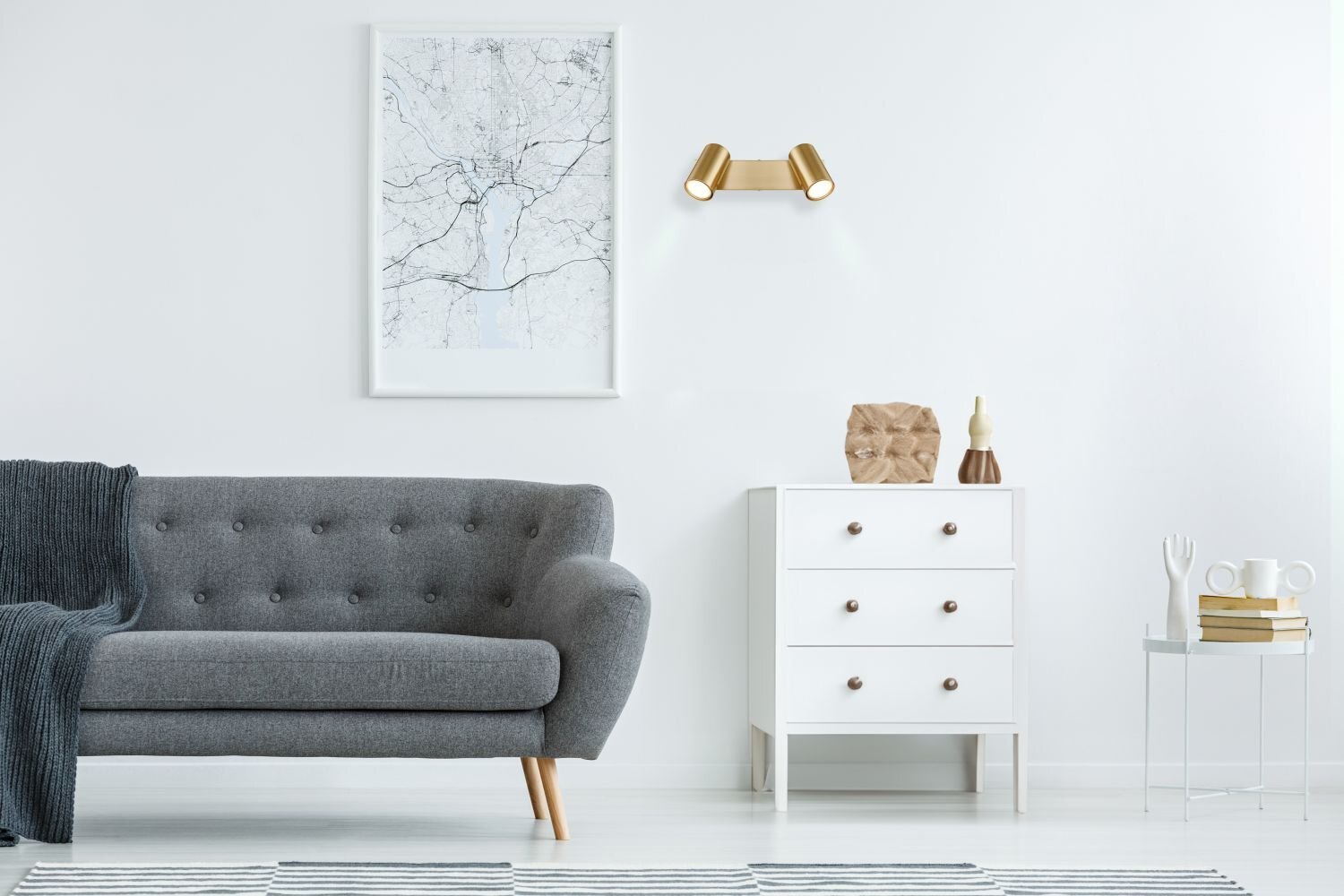 W jasnym wnętrzu znajduje się szara sofa, obok której stoi biała komoda z trzema szufladami. Nad komodą na ścianie zamontowany jest złoty reflektor sufitowy z dwoma punktami świetlnymi.