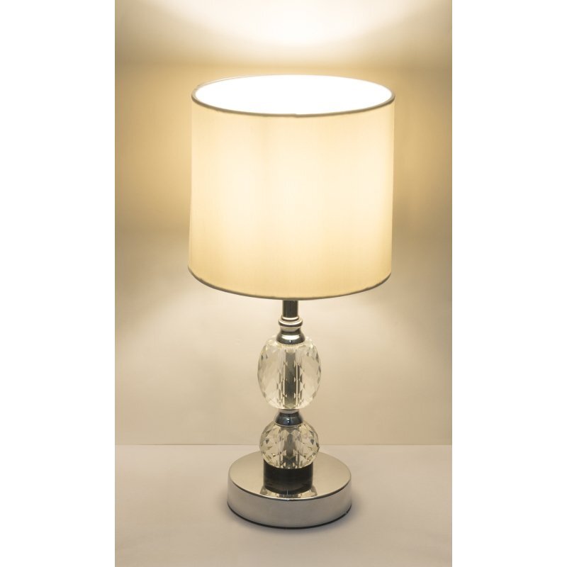 Na obrazie widoczna jest lampa stołowa z białym abażurem i podstawą z chromowanego metalu oraz kryształowego szkła. Lampa emituje ciepłe, rozproszone światło.