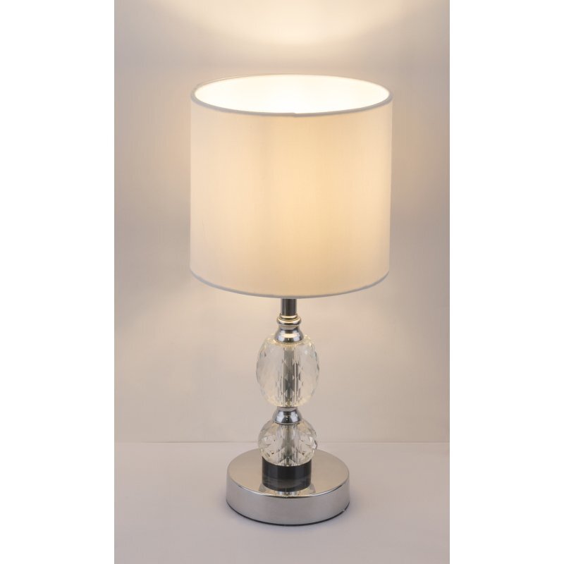 Lampa stołowa z białym abażurem i chromowaną podstawą ozdobioną kryształowymi elementami. Światło jest włączone, co podkreśla elegancki wygląd lampy.