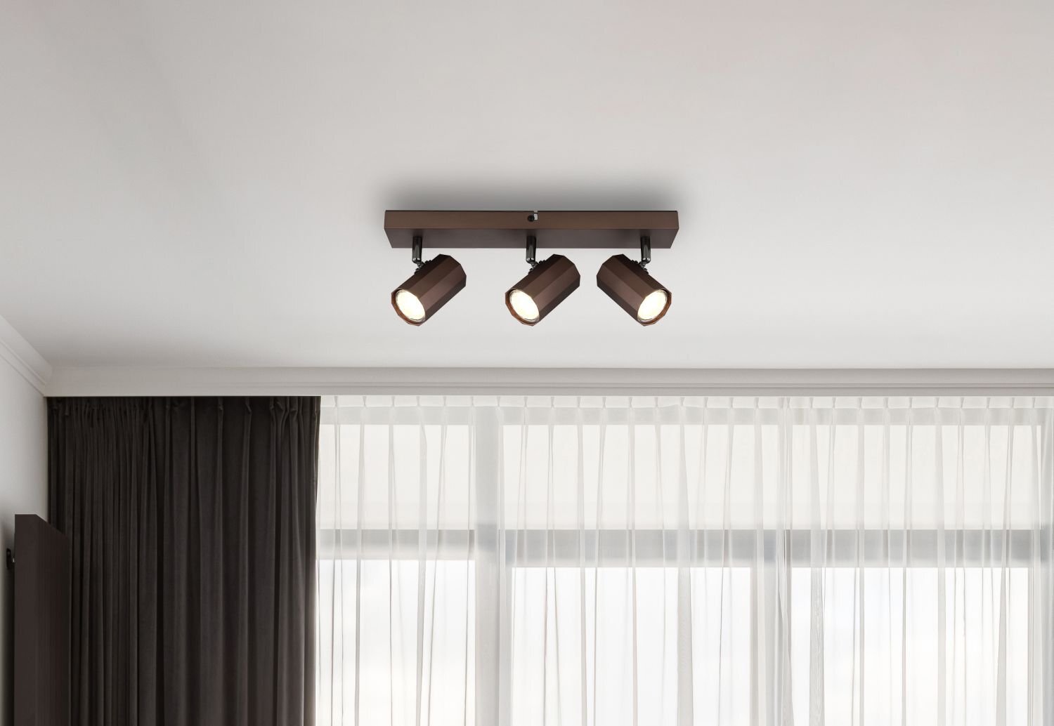 Na suficie zamontowana jest lampa reflektorowa z trzema cylindrycznymi kloszami w ciemnym kolorze. W tle widoczne są zasłony i okno z białą firanką.