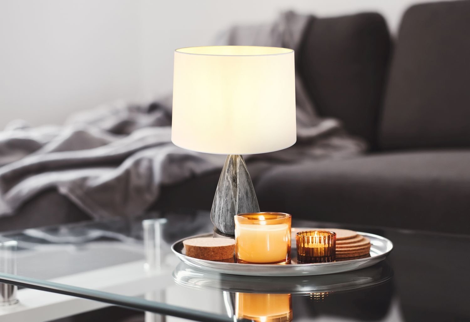 Na szklanym stoliku znajduje się lampa stołowa z jasnym abażurem oraz taca z zapalonymi świecami i podkładkami. W tle widoczna jest ciemna sofa z narzuconym kocem.