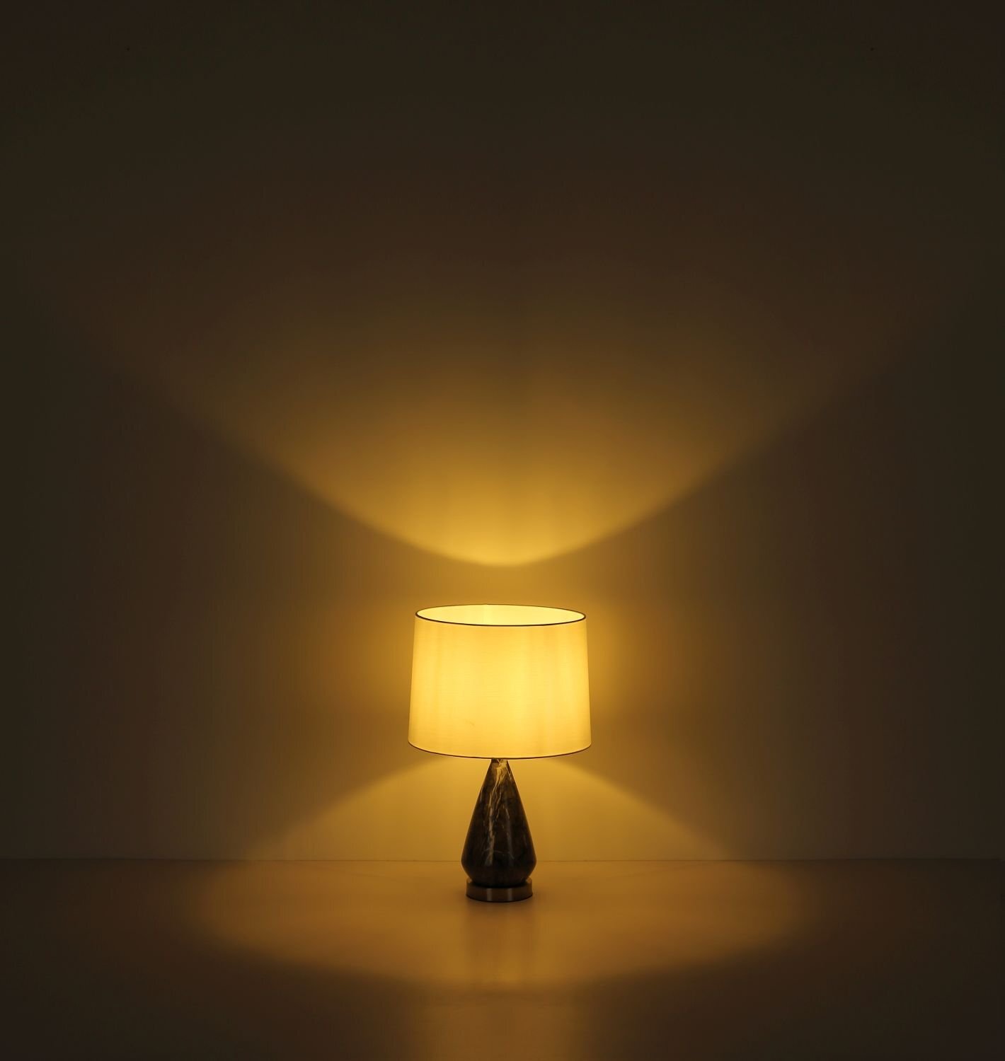 Na obrazie widoczna jest lampa stołowa z jasnym abażurem, emitująca ciepłe, rozproszone światło na ścianę i powierzchnię, na której stoi. Podstawa lampy ma ciemny kolor i stożkowaty kształt.