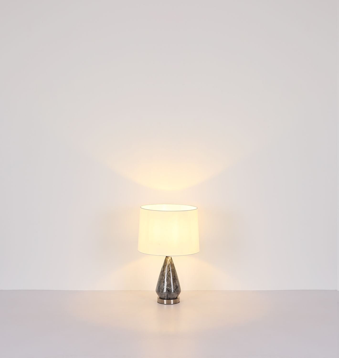Na obrazie widoczna jest lampa stołowa z białym abażurem i podstawą w kształcie stożka o metalicznym wykończeniu. Lampa stoi na jasnej powierzchni, a światło tworzy delikatny wzór na ścianie.
