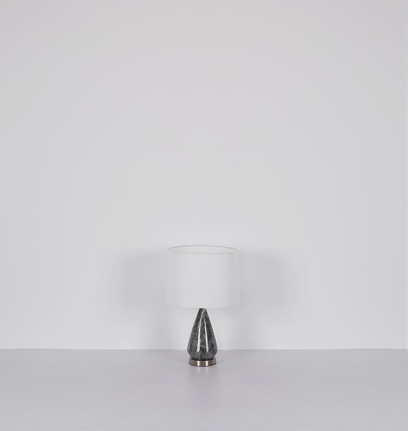 Na obrazie widoczna jest lampa stołowa z białym abażurem i ciemną, geometryczną podstawą. Lampa stoi na jasnej powierzchni, a tło jest jednolicie białe.