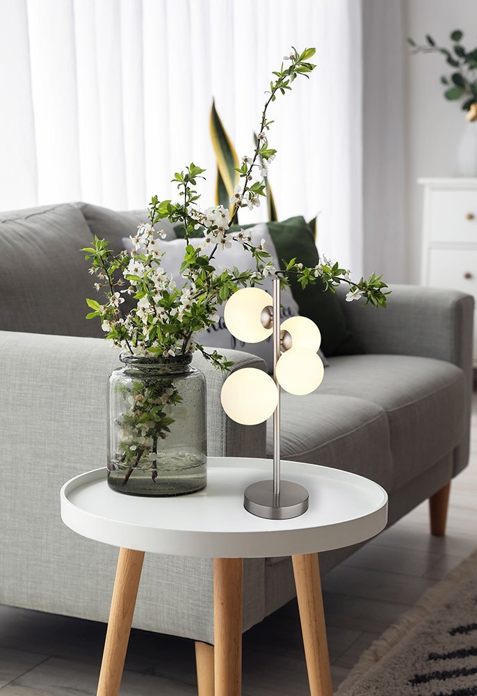 Na okrągłym, białym stoliku stoi lampa stołowa z czterema okrągłymi kloszami oraz wazon z gałązkami kwitnących kwiatów. W tle widoczna jest szara sofa i zasłony.