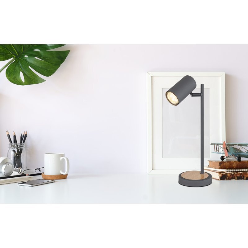 Na białym biurku stoi metalowa lampa stołowa z grafitowym kloszem i drewnianą podstawą. Obok lampy znajduje się kilka książek, model samolotu oraz kubek z przyborami do pisania.