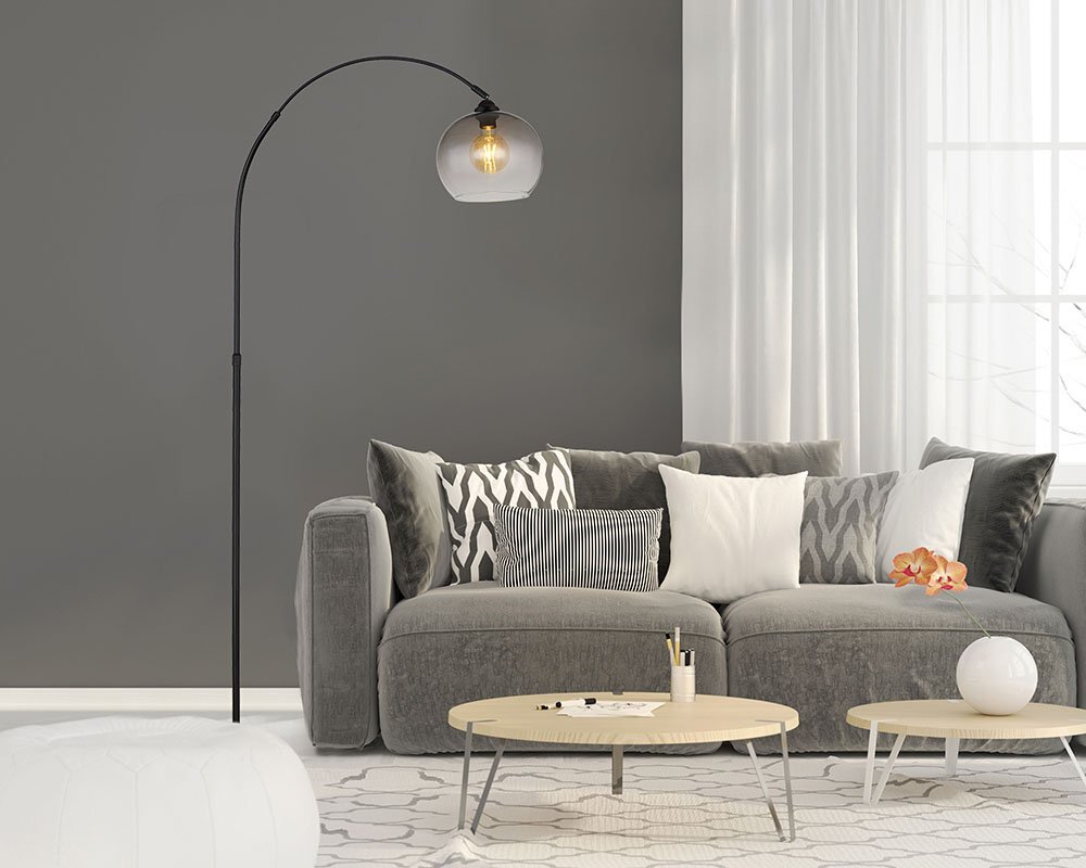 W salonie znajduje się szara sofa z różnorodnymi poduszkami oraz lampa podłogowa z czarnym, zakrzywionym ramieniem i przydymionym kloszem. Na stoliku kawowym leżą drobne przedmioty, a w tle widać zasłony i okno.