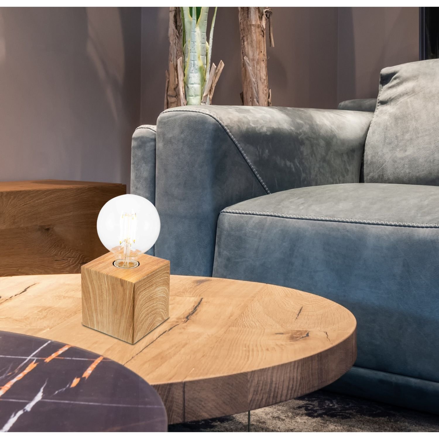 Na drewnianym stoliku znajduje się lampa stołowa z żarówką na drewnianej podstawie. W tle widoczna jest szara, tapicerowana sofa.