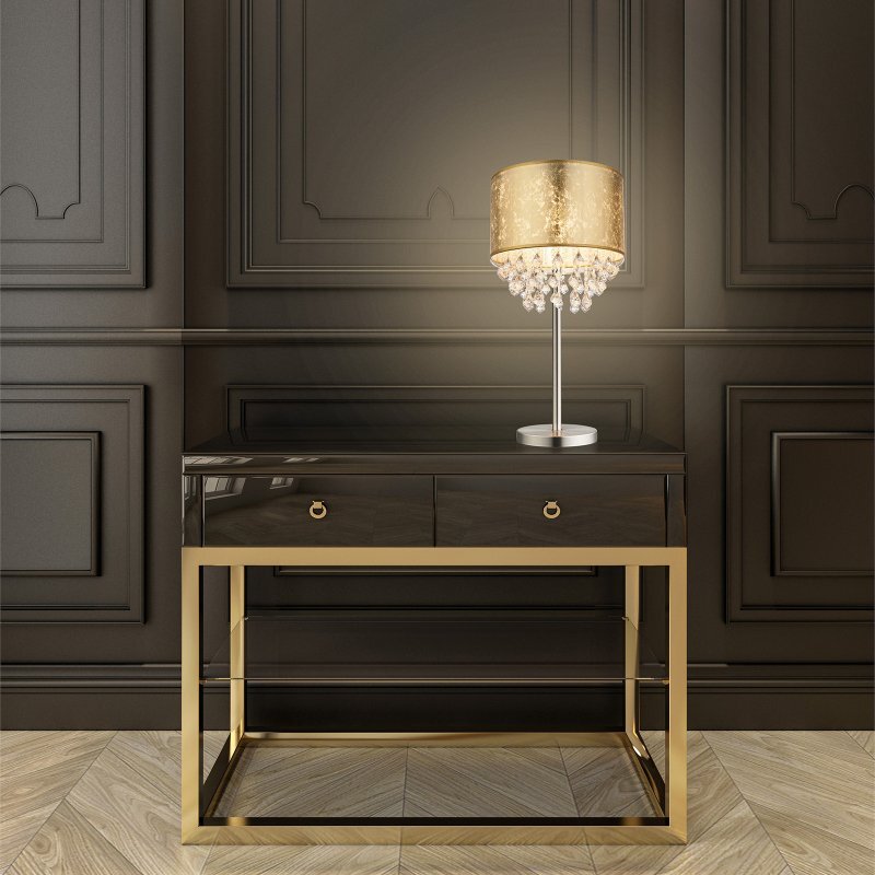 Na obrazie znajduje się elegancka lampa stołowa z satynowym wykończeniem, złotym abażurem i akrylowymi kryształkami, ustawiona na ciemnym stoliku z metalowymi elementami w złotym kolorze. W tle widoczna jest ciemna, ozdobna ściana.