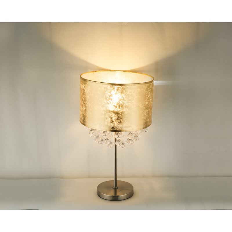 Lampa stołowa z satynowym wykończeniem, posiada złoty abażur i ozdobne kryształki akrylowe. Stoi na metalowej podstawie, a światło rozprasza się przez abażur, tworząc ciepły efekt.
