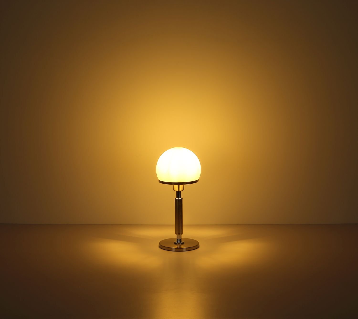 Na obrazie widoczna jest lampa stołowa z okrągłym, mlecznym kloszem emitującym ciepłe, żółte światło. Lampa stoi na metalowej podstawie, a światło tworzy delikatną poświatę na ścianie i powierzchni stołu.