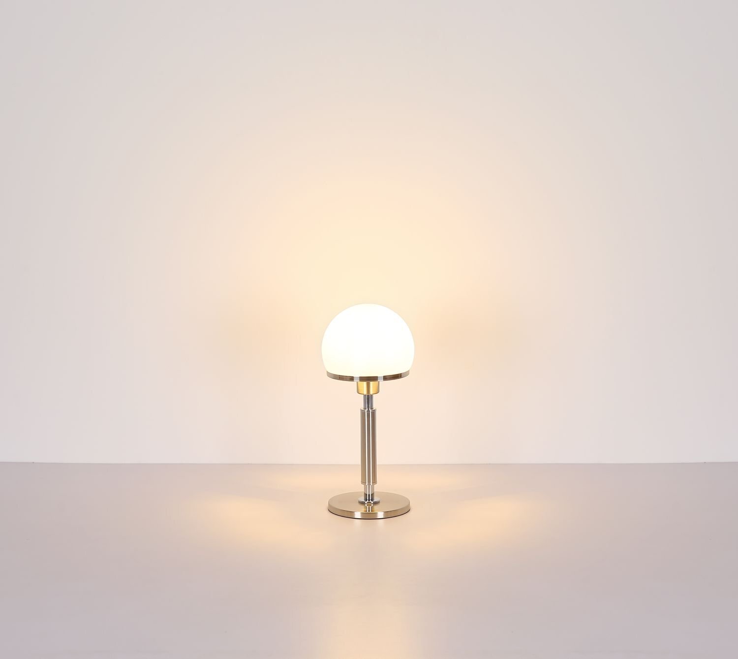Na obrazie widoczna jest lampa stołowa z okrągłym, białym kloszem i metalową podstawą. Lampa emituje ciepłe, rozproszone światło na jasnym tle.