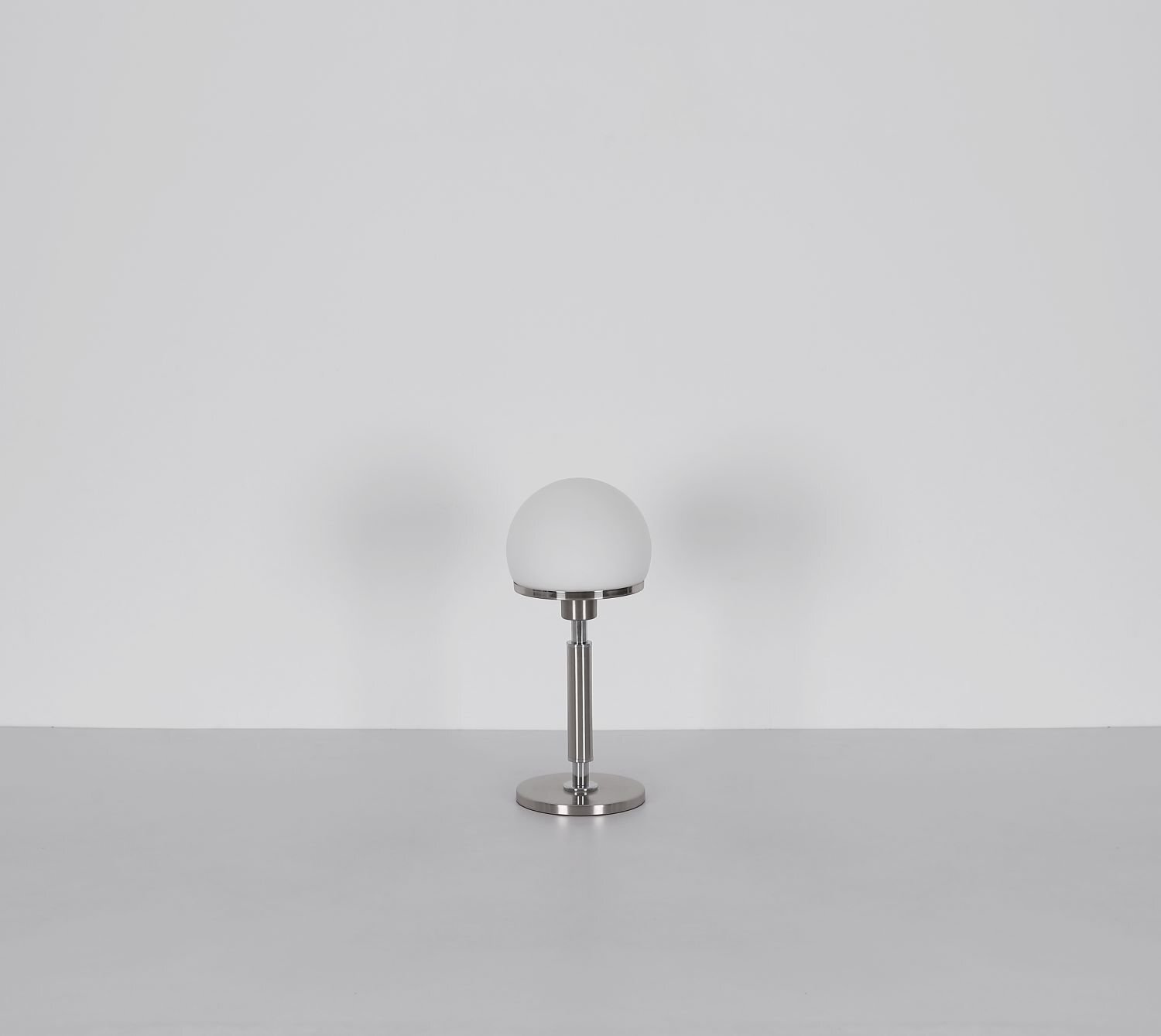 Na obrazie widoczna jest nowoczesna lampa stołowa z białym, kulistym kloszem i metalową podstawą. Lampa stoi na gładkiej powierzchni, a tło jest jednolite i jasne.