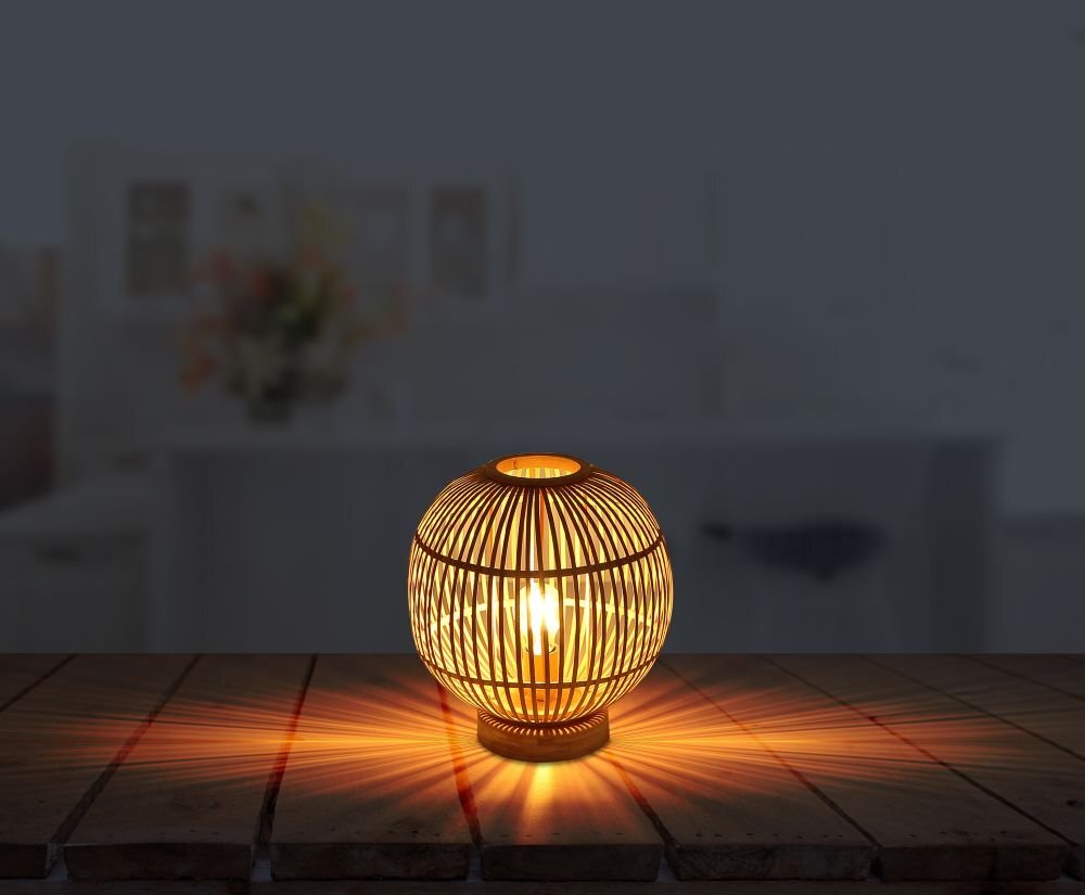 Na drewnianym stole stoi lampa stołowa z naturalnego bambusa, emitująca ciepłe światło. W tle widoczny jest rozmyty obraz wnętrza pomieszczenia.