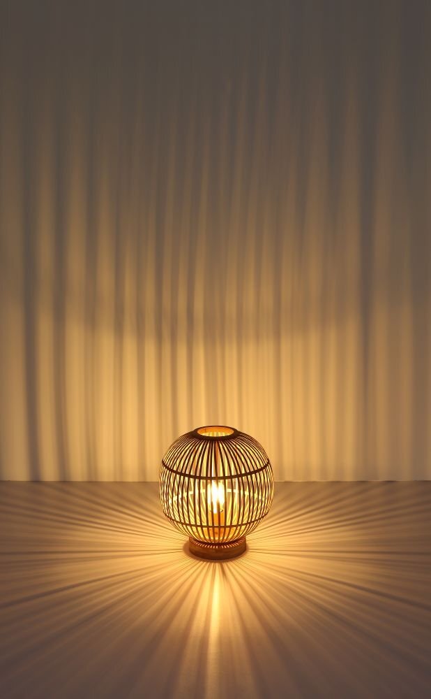 Na obrazie widoczna jest lampa stołowa z naturalnego bambusa, emitująca ciepłe światło, które tworzy wzory na ścianie i powierzchni. Lampa ma kulisty kształt z otwartą strukturą, przez którą prześwituje światło.