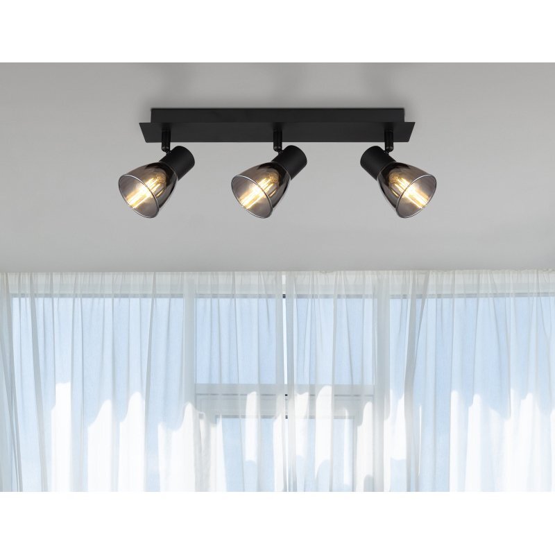 Na obrazie widoczna jest lampa sufitowa z trzema reflektorami w czarnym matowym kolorze, zamontowana na suficie. W tle znajdują się białe, półprzezroczyste zasłony na oknie.