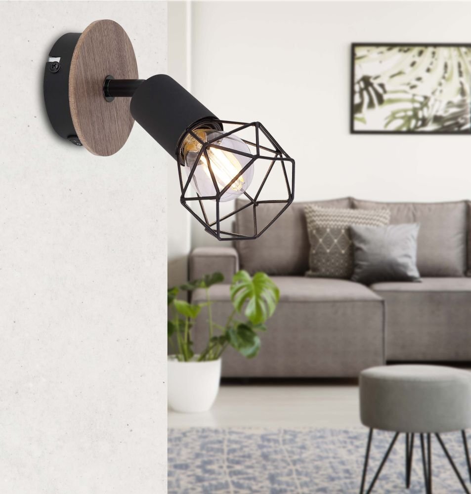 Na obrazie widoczny jest reflektor spot z czarnym metalowym kloszem w geometrycznym kształcie, zamontowany na ścianie z drewnianym elementem. W tle znajduje się szara sofa z poduszkami, roślina w doniczce oraz obraz na ścianie.