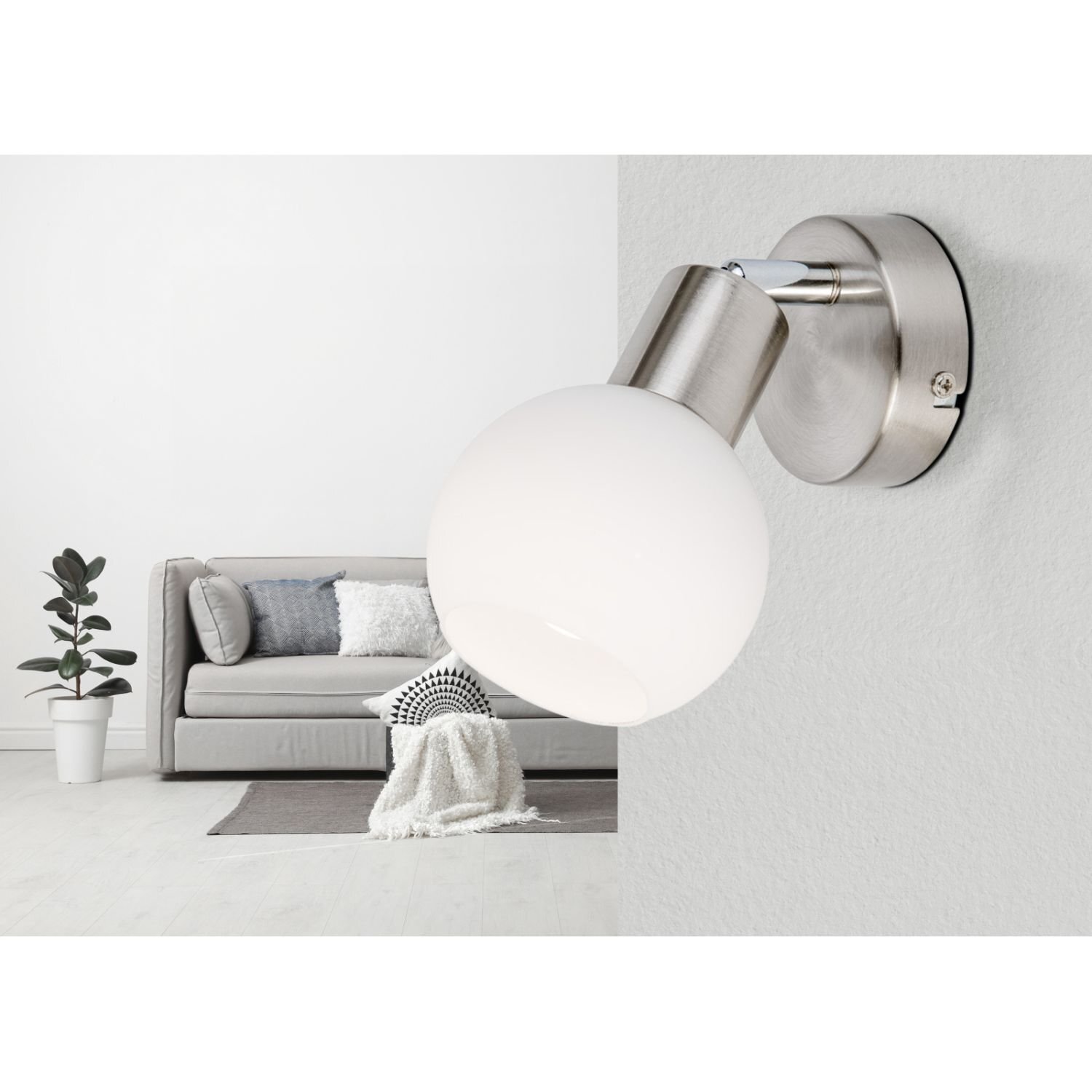 Na obrazie widoczna jest lampa reflektorowa z metalowym uchwytem i białym kloszem zamontowana na ścianie. W tle znajduje się szara sofa z poduszkami oraz roślina w białej doniczce.