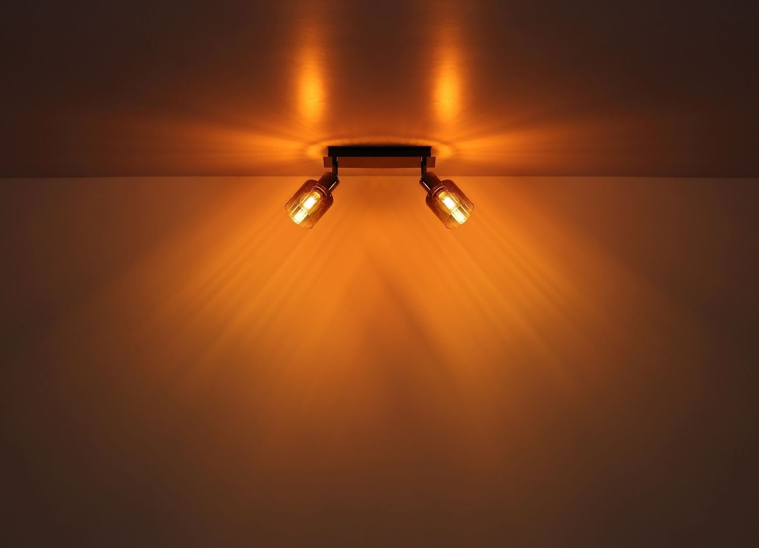 Na suficie zamontowana jest lampa sufitowa z dwoma reflektorami, emitująca ciepłe, pomarańczowe światło, które tworzy promienie na suficie.