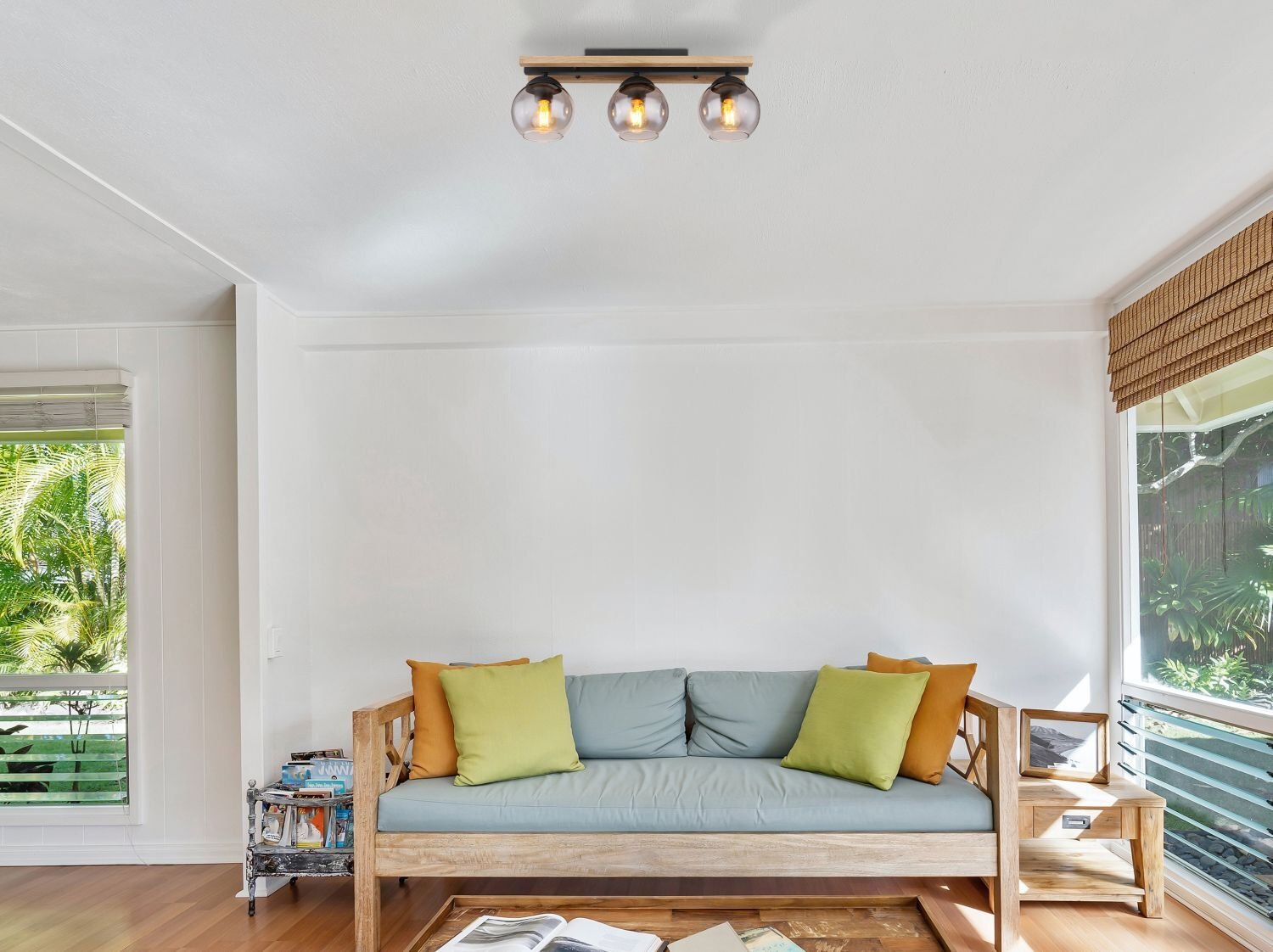 W jasnym pokoju znajduje się drewniana sofa z poduszkami w kolorach zielonym i żółtym. Na suficie zamontowana jest lampa z trzema żarówkami w szklanych kloszach.