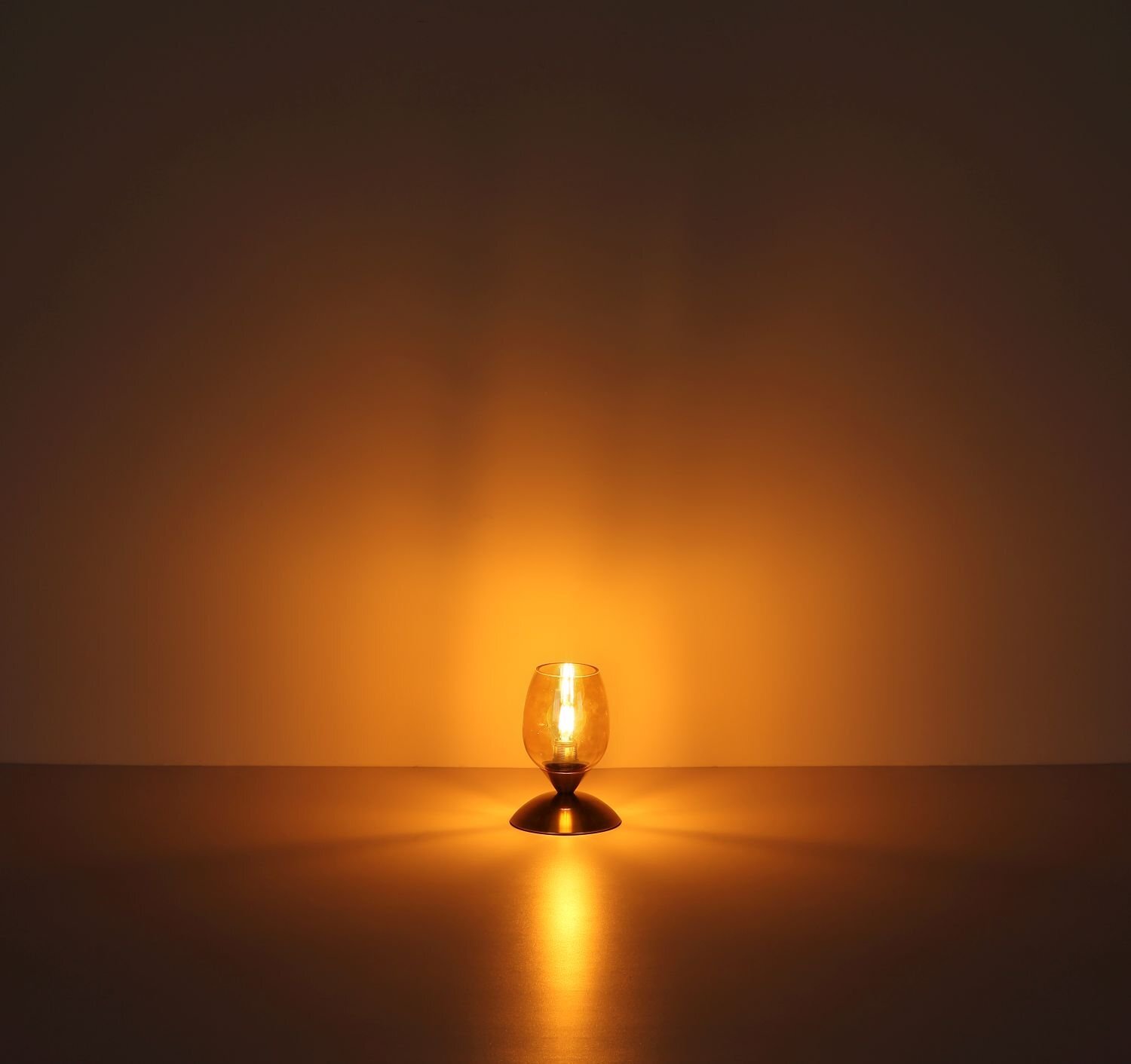 Na obrazie widoczna jest zapalona lampa stołowa o nowoczesnym designie, emitująca ciepłe, żółte światło. Lampa stoi na gładkiej powierzchni, a jej światło tworzy delikatne refleksy na ścianie i podłożu.