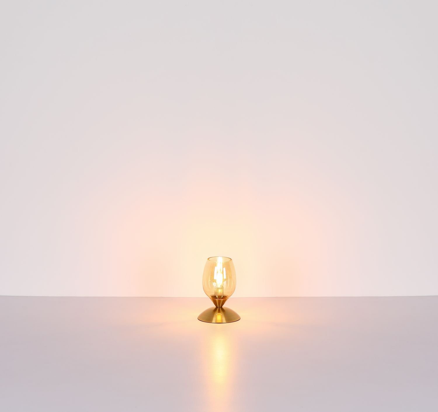 Na obrazie widoczna jest lampa stołowa z przezroczystym kloszem i złotą podstawą, emitująca ciepłe światło na jasnym tle.