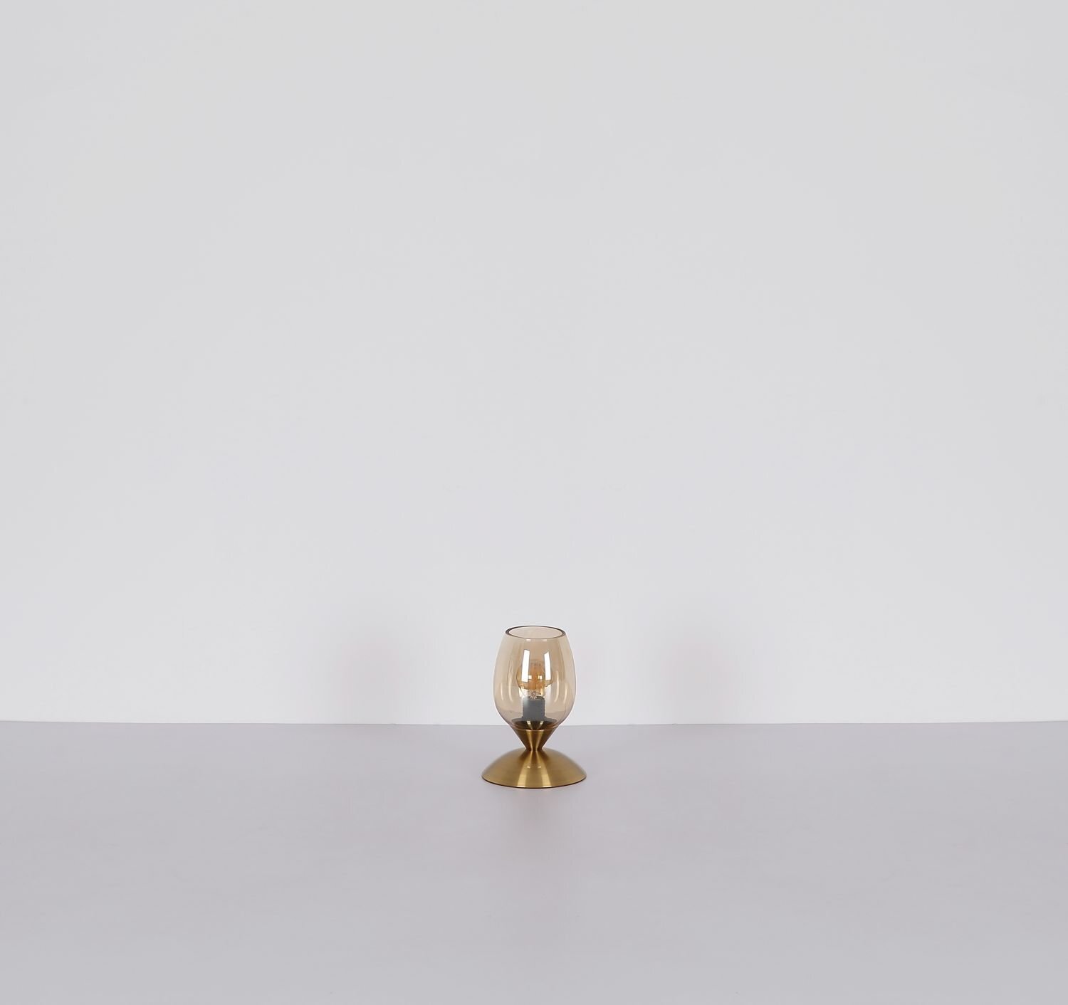 Na obrazie znajduje się lampa stołowa z przezroczystym kloszem i złotą podstawą, ustawiona na jasnym tle.