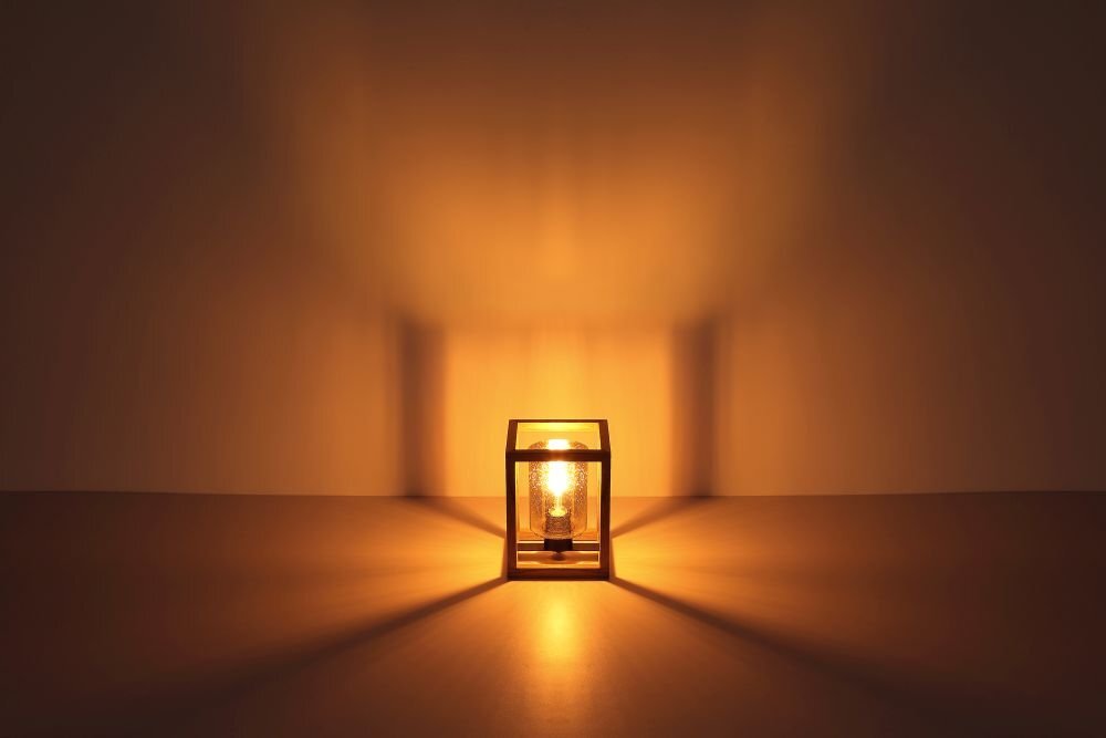 Na obrazie widoczna jest lampa stołowa emitująca ciepłe, żółte światło, umieszczona w przezroczystej obudowie. Światło tworzy symetryczne wzory na powierzchni i ścianie za lampą.