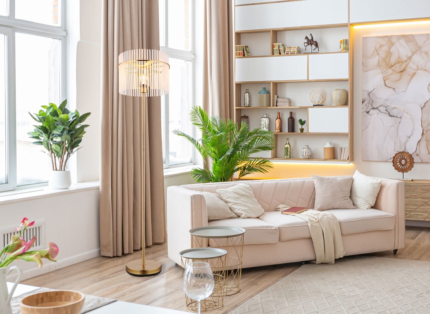 W jasnym, nowoczesnym salonie znajduje się beżowa sofa z poduszkami, obok której stoi stylowa lampa podłogowa z metalowym stelażem i szklanym kloszem. W tle widać regał z dekoracjami oraz duże okna z zasłonami.