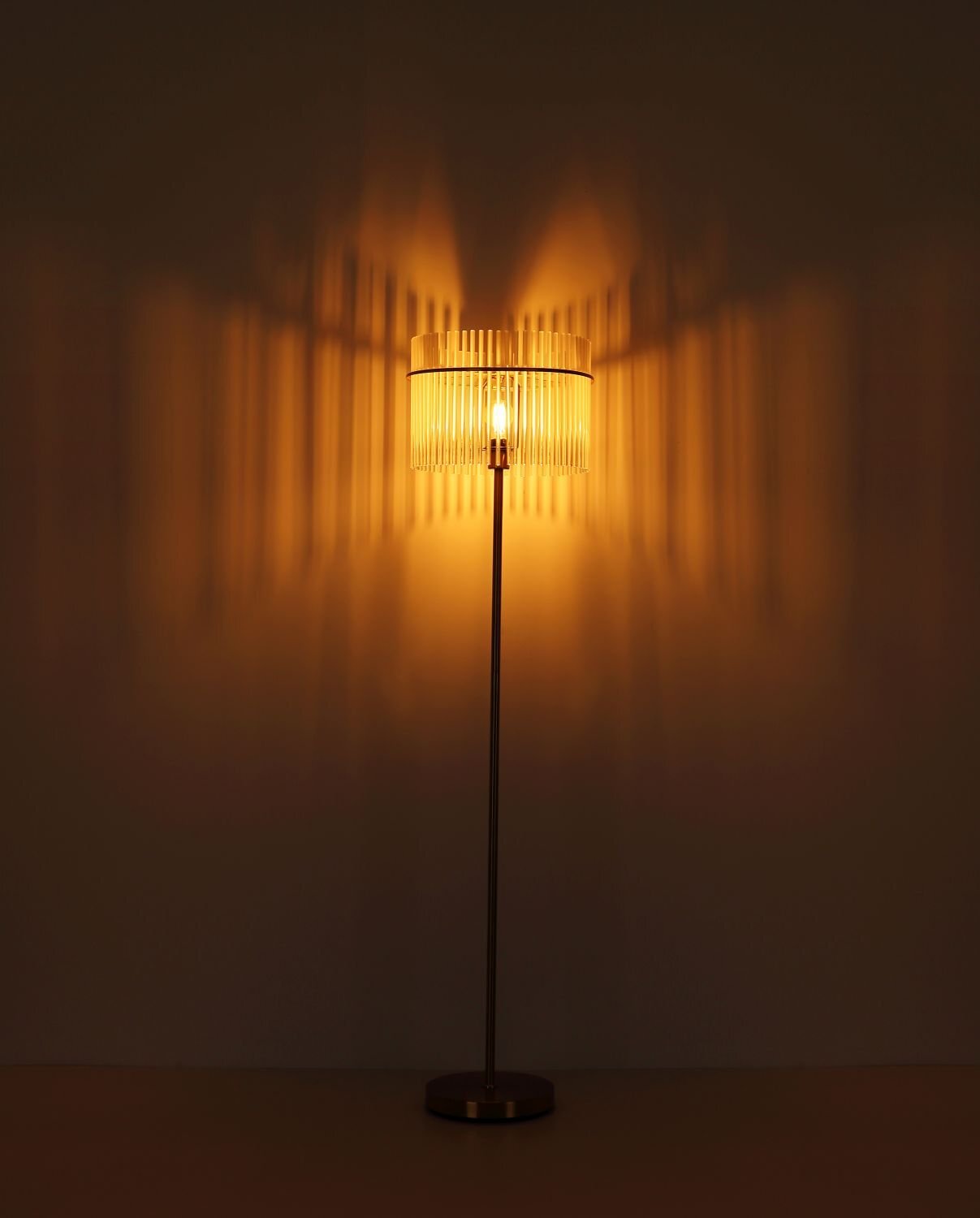 Lampa podłogowa z metalowym stelażem i kloszem, który emituje ciepłe, rozproszone światło, tworząc dekoracyjne wzory na ścianie.