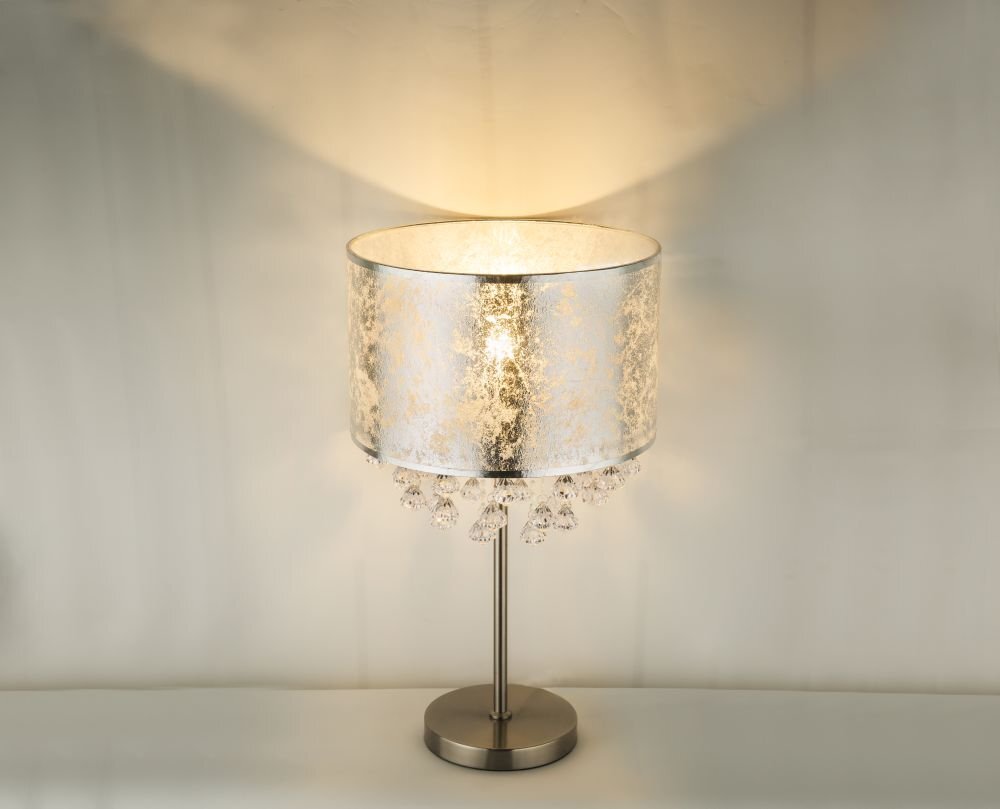 Lampa stołowa z satynowym, srebrnym abażurem tekstylnym i akrylowymi kryształkami. Posiada metalową podstawę i emituje ciepłe światło.