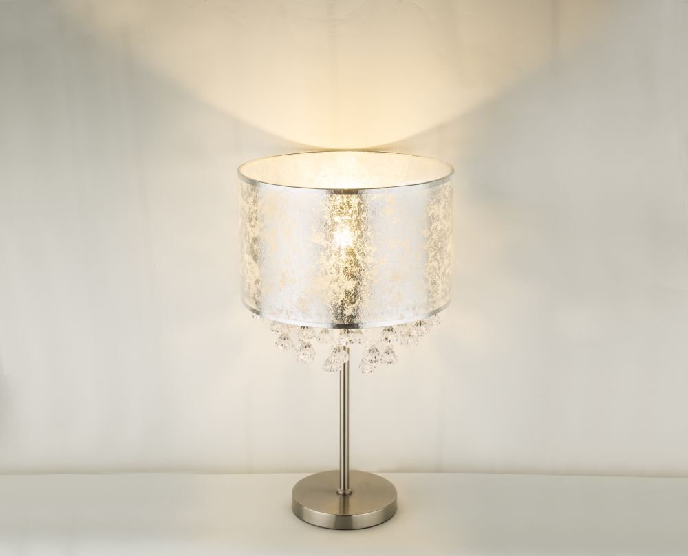 Na obrazie widoczna jest lampa stołowa z satynowym, srebrnym abażurem tekstylnym i akrylowymi kryształkami. Lampa stoi na metalowej podstawie i emituje ciepłe światło.
