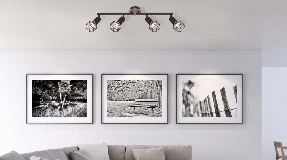 Na suficie zamontowana jest czarna lampa sufitowa z czterema reflektorami, wykonana z metalu i drewna. Na ścianie poniżej znajdują się trzy czarno-białe fotografie w ramkach, przedstawiające krajobrazy.