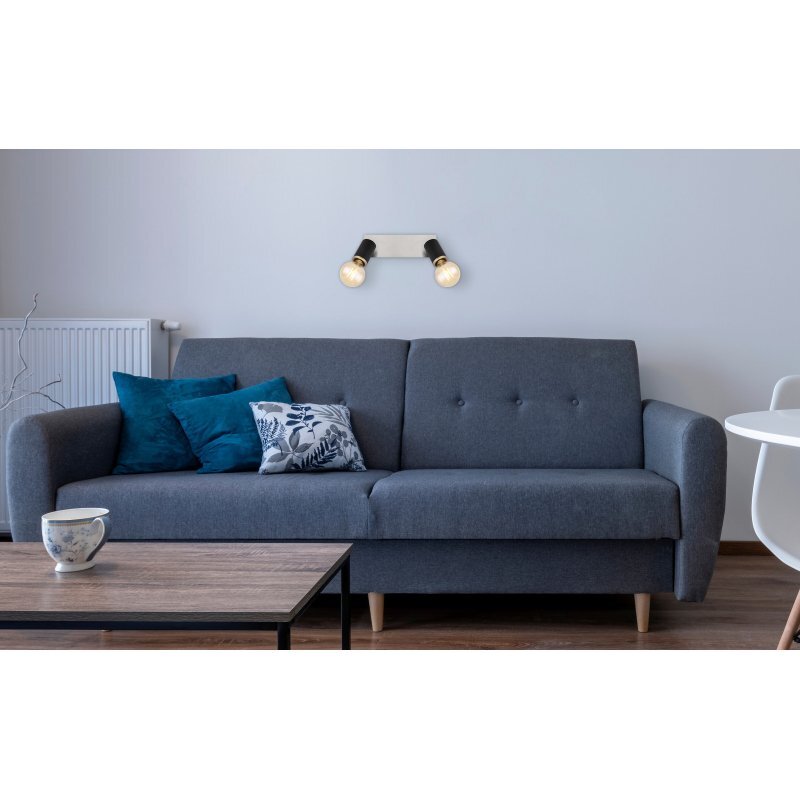 Na ścianie nad szarą sofą zamontowany jest reflektor sufitowy z dwoma żarówkami. Sofa ozdobiona jest poduszkami w odcieniach niebieskiego i wzorzystymi.