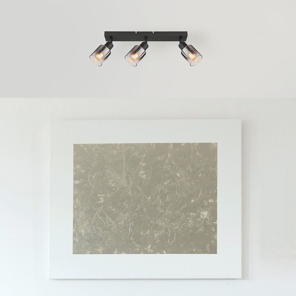 Na suficie zamontowana jest lampa sufitowa z trzema reflektorami w czarnym matowym wykończeniu i przydymionym szkle. Poniżej znajduje się obraz w białej ramie z abstrakcyjnym wzorem.