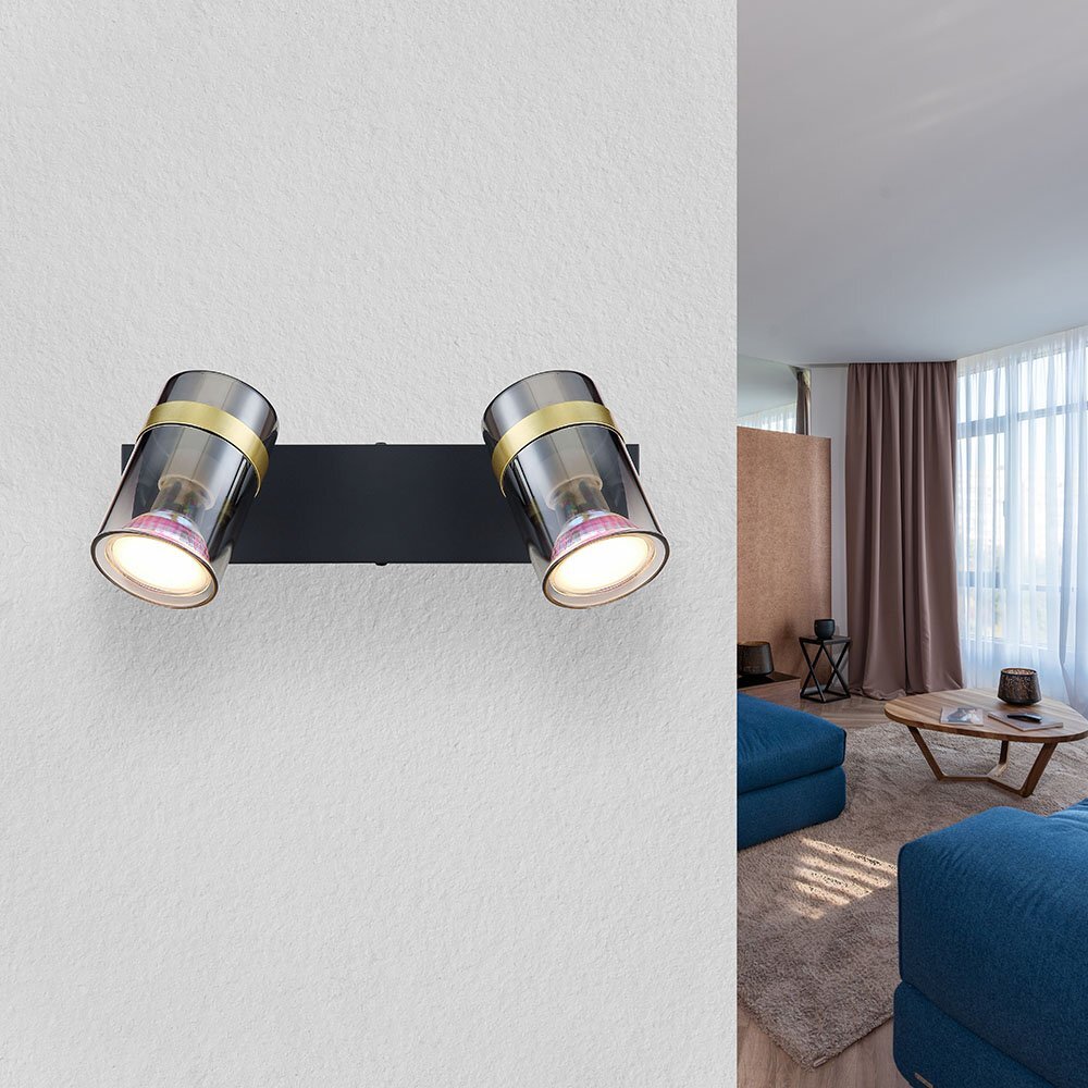 Lampa sufitowa z dwoma reflektorami w czarnym matowym wykończeniu, z przydymionym szkłem i mosiężnymi elementami, zamontowana na ścianie. W tle widoczny jest fragment salonu z niebieskimi sofami i dużym oknem.