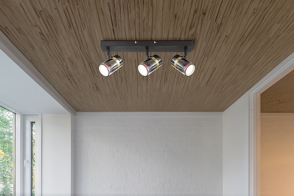 Na suficie zamontowana jest lampa sufitowa z trzema reflektorami w czarnym matowym wykończeniu z przydymionym szkłem i mosiężnymi elementami. Pomieszczenie ma drewniany sufit i białe ceglane ściany.