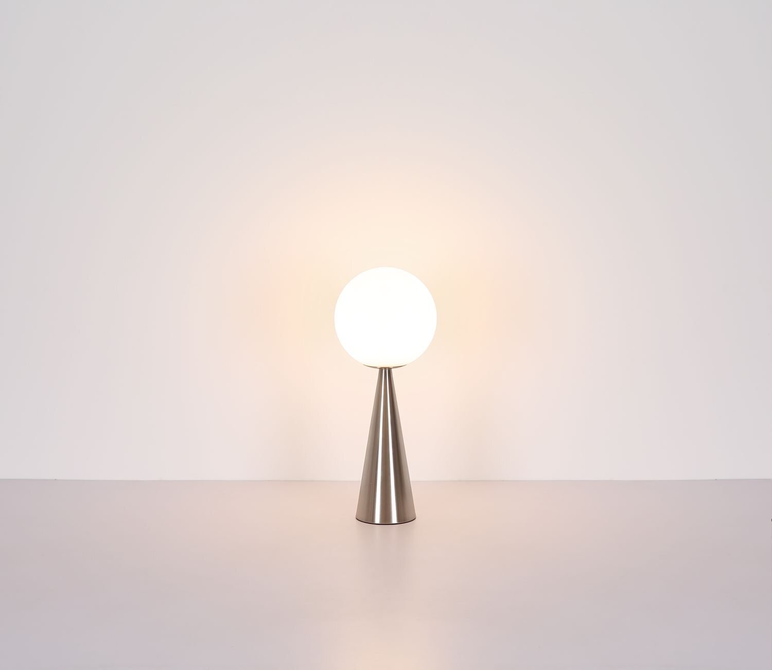 Na obrazie widoczna jest lampa stołowa z kulistym kloszem na stożkowej podstawie w metalicznym kolorze. Lampa emituje ciepłe, rozproszone światło na jasnym tle.