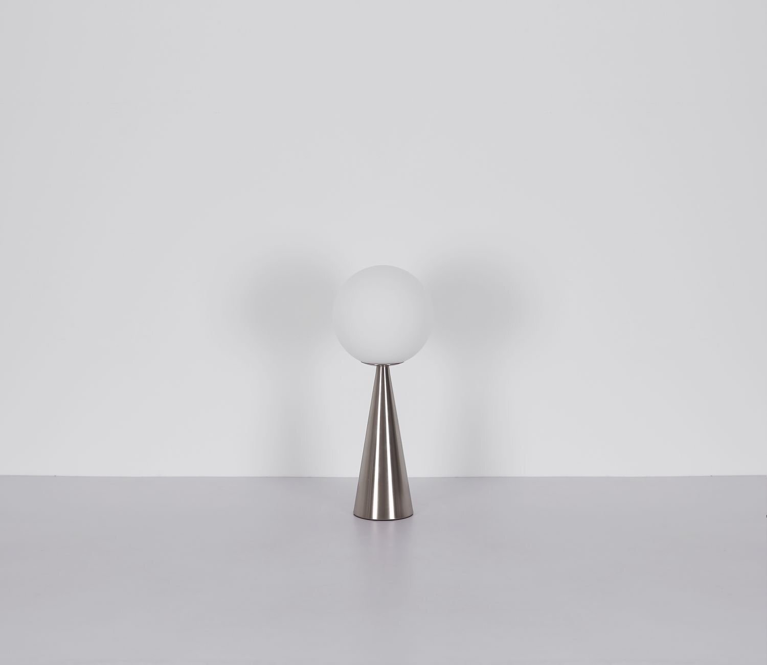 Na obrazie widoczna jest lampa stołowa z białym, kulistym kloszem osadzonym na stożkowej, metalicznej podstawie. Tło jest jednolite i jasne.