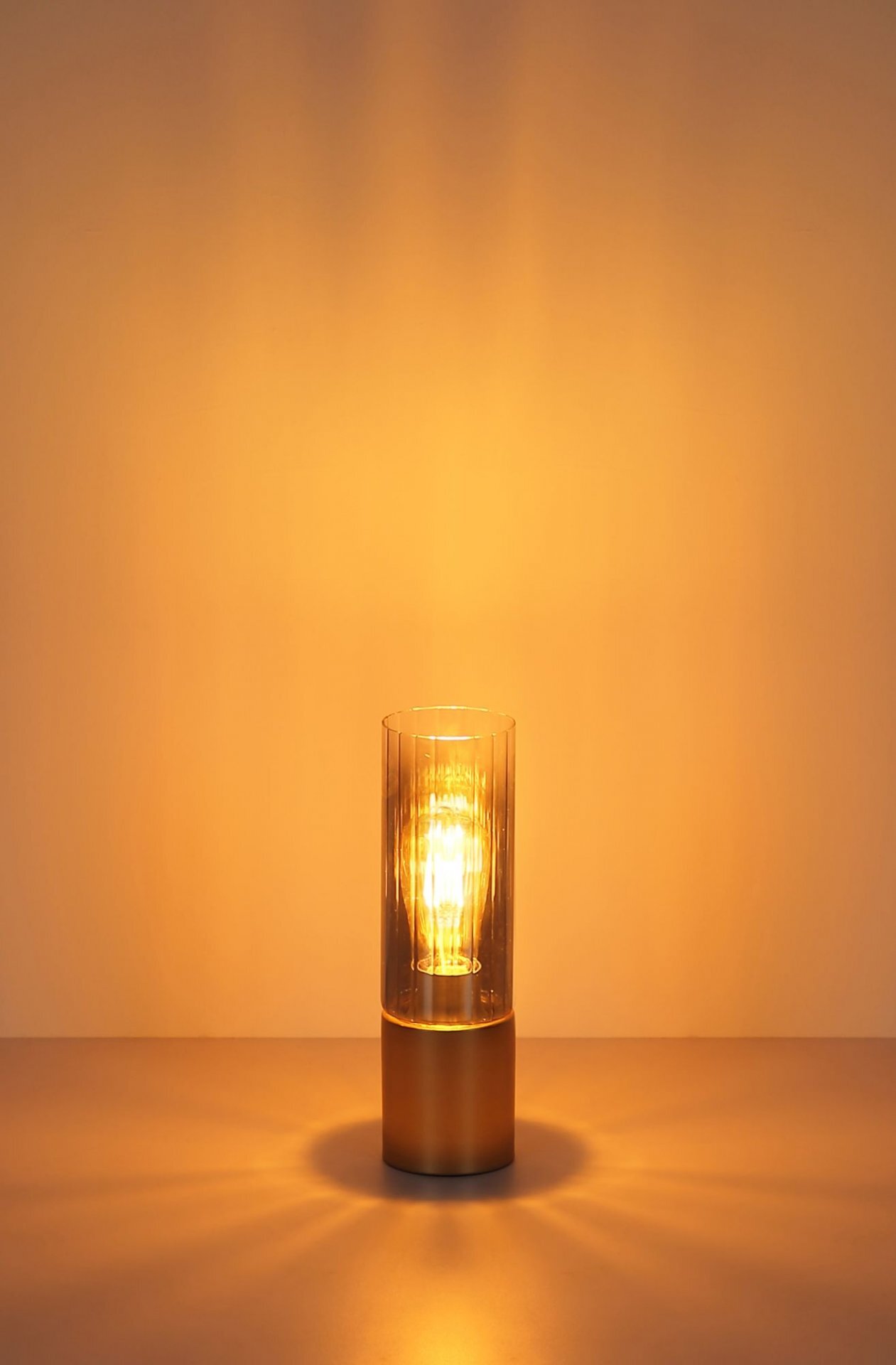 Na obrazie widoczna jest lampa stołowa z przezroczystym kloszem, emitująca ciepłe, żółte światło. Lampa stoi na gładkiej powierzchni, a światło tworzy delikatne refleksy na tle.