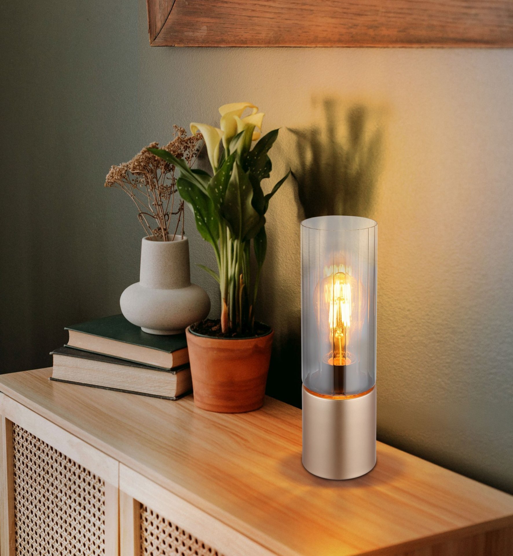 Na drewnianej komodzie stoi szklana lampa stołowa z widoczną żarówką, obok której znajdują się doniczki z roślinami i stos książek. Ściana w tle jest w ciepłym odcieniu, a na niej wisi drewniana rama.