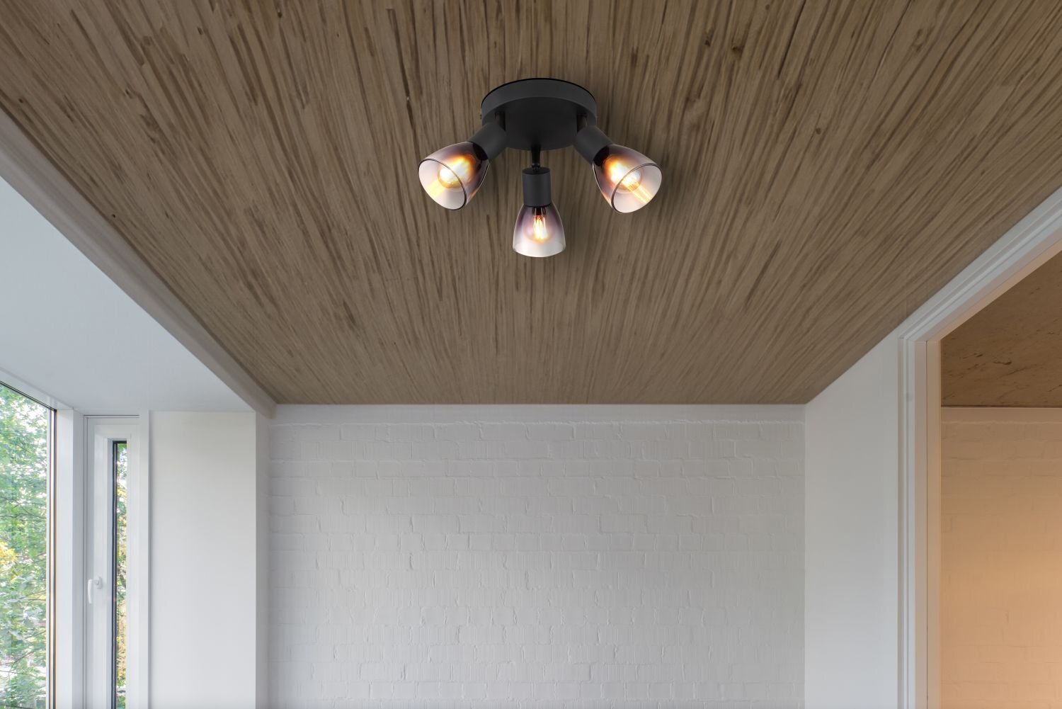 Na suficie z drewnianymi panelami zamontowana jest czarna lampa sufitowa z czterema reflektorami. W tle widoczna jest biała ceglana ściana i okno z widokiem na zewnątrz.