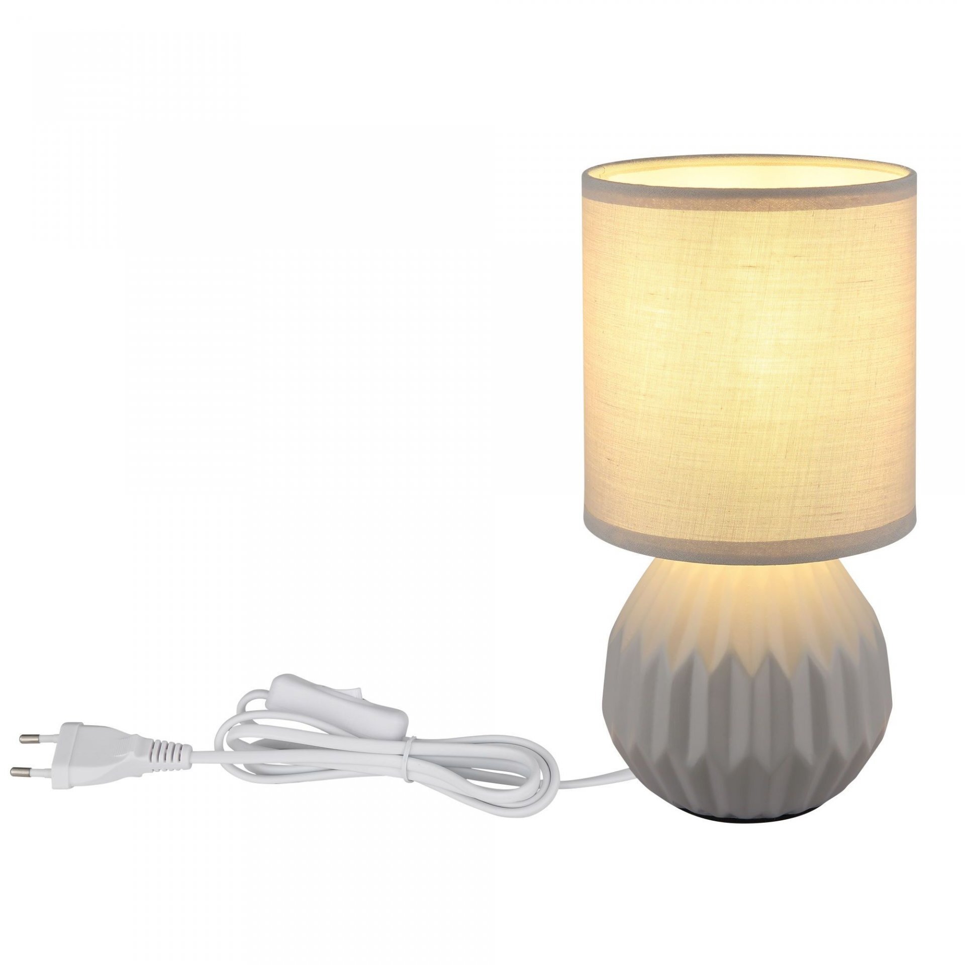Na obrazie widoczna jest lampa stołowa z jasnym abażurem i żłobioną, szarą podstawą. Obok lampy znajduje się biały przewód z wtyczką.