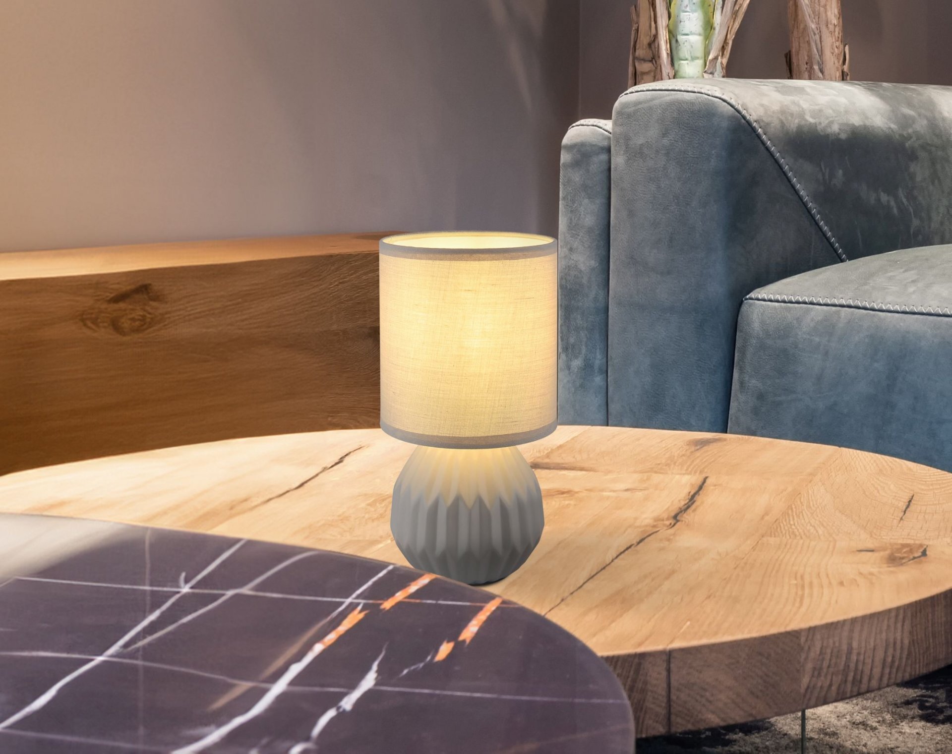 Na drewnianym stoliku znajduje się elegancka lampa stołowa z jasnym abażurem i ozdobną podstawą. W tle widoczna jest część kanapy w kolorze szarym.