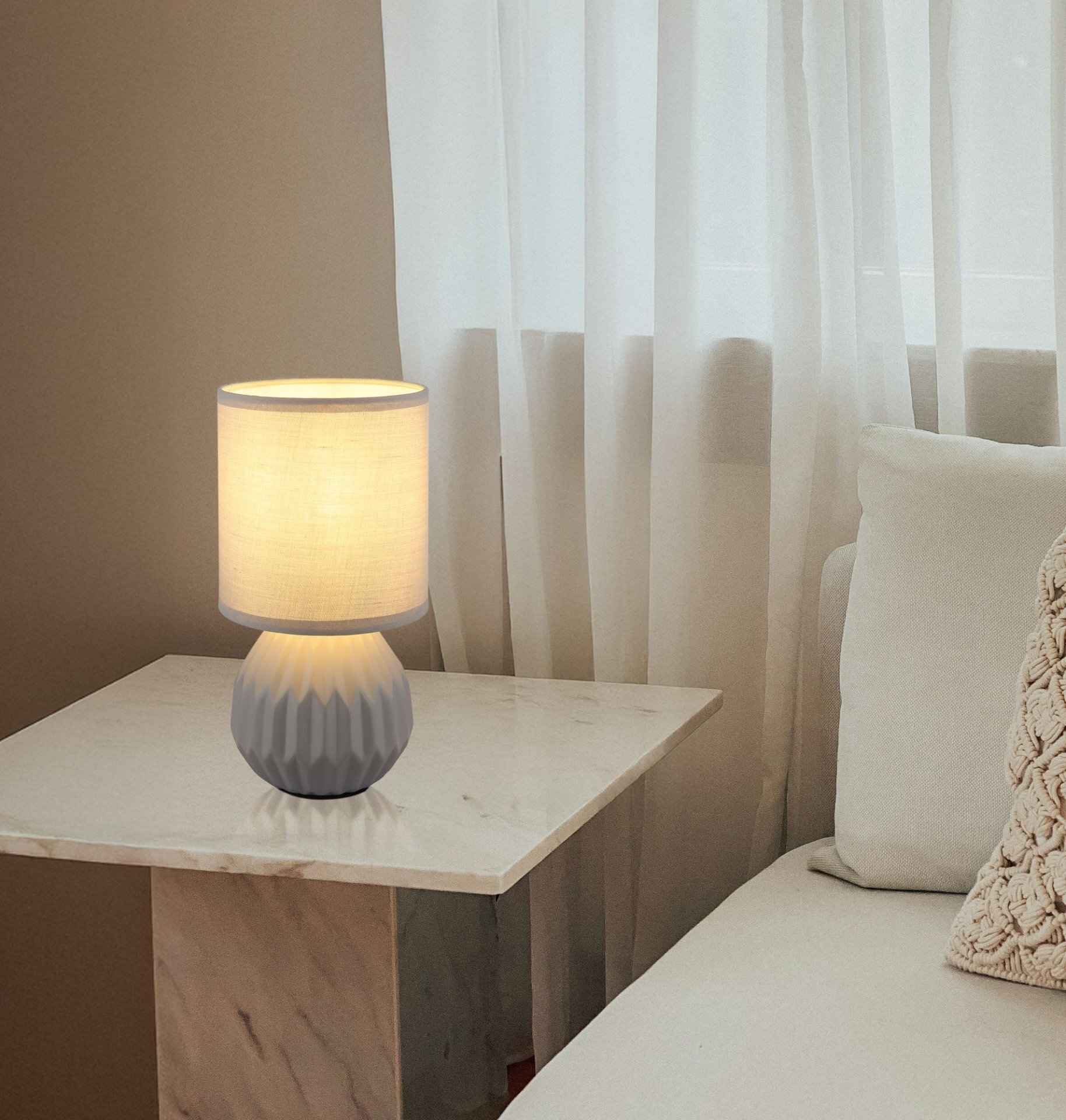 Na marmurowym stoliku znajduje się lampa stołowa z jasnym abażurem i szarym, żłobionym podstawą. W tle widoczne są białe zasłony i fragment jasnej kanapy z poduszką.