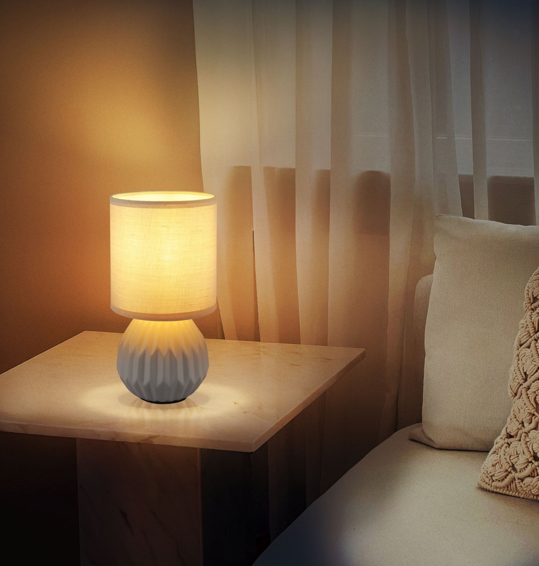 Na stoliku znajduje się włączona lampa stołowa z jasnym abażurem i ozdobną podstawą. W tle widoczne są zasłony i fragment kanapy z poduszką.