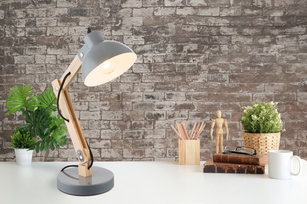 Na białym biurku stoi lampa biurkowa z drewnianym wysięgnikiem i szarym kloszem, obok której znajdują się rośliny w doniczkach, drewniany manekin, kubek oraz książki. W tle widoczna jest ściana z cegieł.