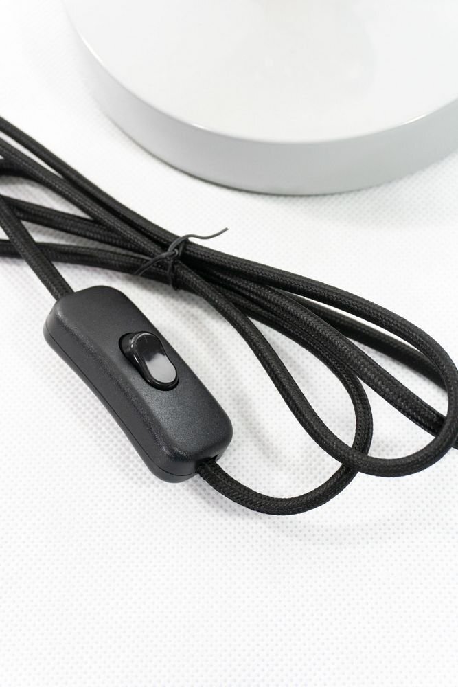 Czarny kabel z wyłącznikiem leży na białej powierzchni obok białej podstawy lampy.